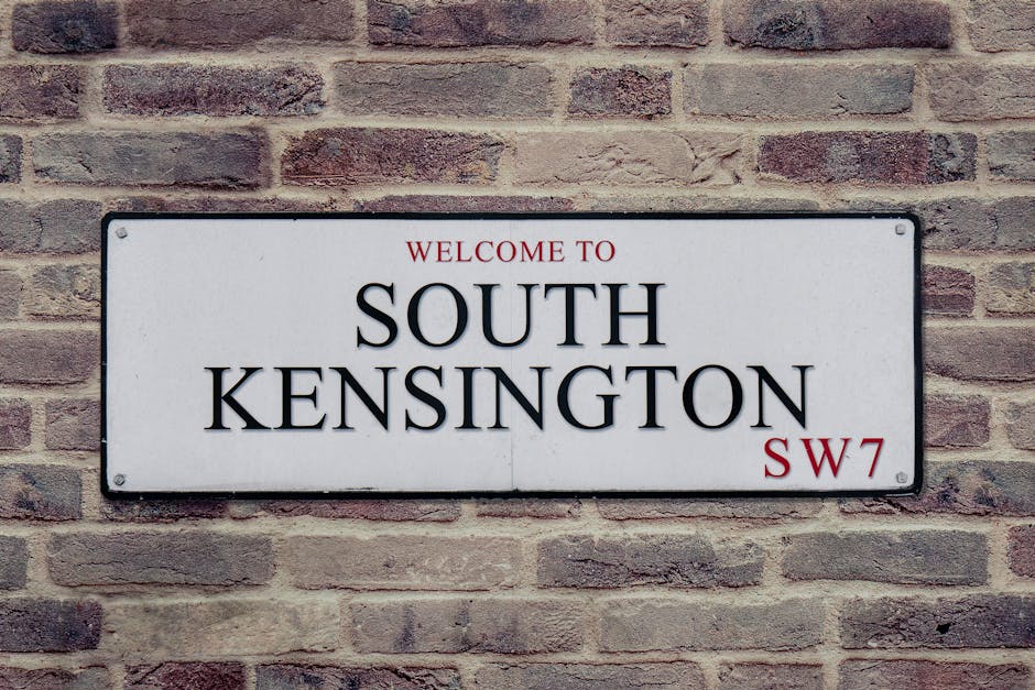 A rectangular metal street sign mounted on a red brick wall, reading 'Welcome to South Kensington SW7' in black and red text. The sign has a white background with slightly rounded edges, and the brick